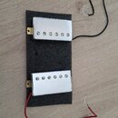 Dvě elektrické kytarové snímače s chromovaným krytem, které jsou připevněny k šedému pěnovému podkladu. Snímače mají pět viditelných šroubů v řadě. Z…