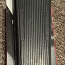 Černý sustain pedál s žebrovaným povrchem, umístěný na bílé krabici. Na pedálu jsou vyrazeny nápisy INSTRUMENT a AMPLIFIER. Povrch plastu vykazuje mí…