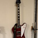 Kytara s červeným trupem a bílým pickguardem, která visí na stěně. Na hlavě hředu je bílý nápis Gibson. Na bílém pickguardu je vidět červený nápis a…