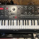 Černý syntetizátor s klaviaturaí a nápisem Roland JDXi. Přední panel obsahuje množství ovládacích knoflíků, tlačítek a displej s červeným textem. V p…