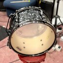Kytarový snare drum s černým mramorovaným povrchem a chromovanými obručemi. Na spodní bláně je vidět logo TAMA a nečitelné nápisy v horní části. Nást…