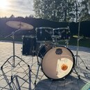 Černá bubnová sada s chromovaným kováním, zahrnující basový buben, dva tom-tomy, floor tom a jeden činel. Na přední bláně basového bubnu je logo MAPE…