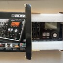 Na fotografii je digitální nahrávací studio Boss BR-800 v bílém polystyrenovém obalu a vedle něj jeho originální krabice. Zařízení je černé s ovládac…