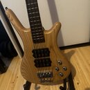 Světle hnědá elektrická basová kytara s přírodným dřevem a viditelnou masivní strukturou zrna. Nástroj má dva černé humbuckery, čtyři ovládací knoflí…