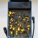 Otevřený šedý kovový box s vnitřní elektronickou deskou a černým plastovým blokem ve horní části. Na desce jsou osazeny různé součástky, včetně integ…