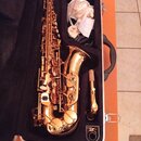 V otevřeném černém kufru leží saxofon s lesklým, růžozlatým povrchem. Vedle nástroje je umístěn mouthpiece s troškou rýžového rýdu a bílá látka. Na n…