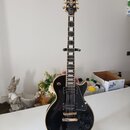 Černá elektrická kytara typu Les Paul s bílými hrankami a zlatým hardwarem. Na hlavě je nápis Epiphone a logo v podobě křídla. Kytara má dva humbucke…