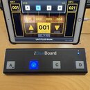 Fotografie zobrazuje černý MIDI kontrolér iRig BlueBoard s čtyřmi bílými tlačítky označenými písmeny A, B, C a D. Tlačítko B je podsvícené modře a v…