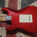 Zadní strana červené elektrické kytary s tvarem typu Stratocaster. Na zadní straně těla je bílý plastový kryt elektroniky připevněný šesti šrouby. Na…