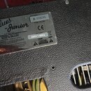 Detail kovového štítku na zadní straně kytarového zesilovače s černým texturovaným povrchem. Na štítku je uveden název Blues-Junior a nápis A PRODUCT…