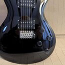 Černá elektrická kytara s lesklým povrchem. Nástroj má dva humbuckery, chromovaný most a čtyři černé ovládací knoflíky. Na povrchu nejsou viditelné ž…