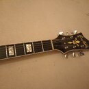 Detail krku a hlavy elektrické kytary s černým povrchem. Na hlavě je zlatý nápis "Gibson" a zlatý symbol v podobě stylizovaného písmena Y. Hmatník je…
