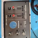 Šedý USB audio interface s černým rámem. Na přední straně jsou nápisy Solid State Logic a SSL 2+. Zařízení obsahuje dva kanály s ovládacími prvky GAI…