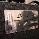 Detail kovového štítku na zadní straně černého zesilovače s texturovaným povrchem. Na štítku je nápis Deluxe a text A PRODUCT OF FENDER MUSICAL INSTR…