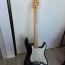 Černá elektrická kytara s bílým pickguardem a hnedým javorovým hridlem. Na hlavě je viditelné logo Fender a nápis Stratocaster. Nástroj vykazuje znám…