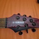 Hlava elektrické kytary v purpurové barvě s viditelnou strukturou dřeva. Na povrchu je bílý nápis YAMAHA s doprovodným logem. Jsou vidět šest ladicíc…