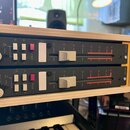 Dvě identické rackové jednotky v černém provedení umístěné v dřevěném rámu. Každá jednotka obsahuje stříbrný otočný knoflík, čtyři bílá tlačítka, jed…