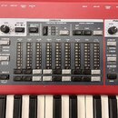 Červený elektronický klavír s ovládacím panelem označeným nápisy ORGAN, PIANO a PROGRAM. Panel obsahuje řadu přepínačů pro různé registry (např. BASS…