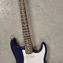 Tmavě modrá elektrická kytara s bílým pickguardem a hnědým hmatníkem. Na hlavě je viditelné logo Stagg. Kytara má tři snímače typu single-coil, pětip…