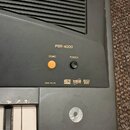 Černý digitální klavír s nápisem PSR-4000. Na ovládacím panelu jsou tlačítka DEMO a POWER, žlutá kontrolka a nápisy DISK IN USE, GENERAL MIDI, ORCHES…