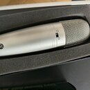 Stříbrný kondenzátorový mikrofon s kovovou síťkovou hlavou, umístěný v černé pěnové výplni přepravního kufru. Na těle mikrofonu je vidět symbol CE a…