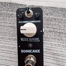 Černý kytarový efekt s bílým velkým otočným knoflíkem uprostřed. Na šasi jsou nápisy RUDE MOUSE a SONICAKE, dále popisky LEVEL, GAIN, FILTER a přepín…