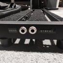 Černý kovový pedalboard s povrchem z černého plsti. Na přední hraně jsou dva zapuštěné konektory a nápisy MONO a STEREO. Nástroj stojí na čtyřech čer…