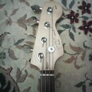 Hlava basové kytary z jasného dřeva s čtyřmi ladicími kolíky. Na hlavě je černý nápis Squier J BASS a pod ním menší nápis Fender. Povrch dřeva vykazu…