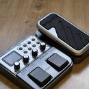 Šedý kytarový procesor s ovládacími knoflíky, displejem a dvěma černými spínacími pedály. Na přístroji jsou viditelné nápisy MG-100, Modeling Guitar…