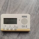 Bílý digitální ladič s žlutým spodním okrajem. Na přístroji je nápis KORG a model CA-2 CHROMATIC TUNER. Displej zobrazuje hodnotu 440, pod ním jsou t…