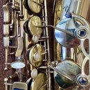 Detailní záběr na zlatavý kovový saxofon s perleťovými klapky. V horní části je vidět nápis MADE IN FRANCE a logo s textem Selmer Paris Action Serie…