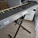 Šedý digitální klavír značky KORG, model N264, umístěný na černém X-stojanu. Na boku je viditelný štítek s nápisy KORG, MODEL N264 a SERIAL No. 00332…