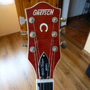 Detail hlavy červené kytary s viditnou strukturou dřeva. V horní části je bílý nápis GRETSCH a pod ním bílé logo ve tvaru podkovy. Na spodní části hl…