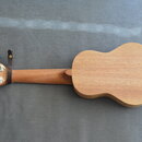 Fotografie zachicuje zadní stranu dřevěné ukulele v přírodní barvě. Na hmatníku je u hlavy nasazený hnědý kožený pásek. Nástroj nemá žádné viditelné…