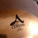 Detail zlatavého činchového činely s viditelným broušením povrchu. Na činely je černý nápis CUSTOM s logem v podobě stylizovaného písmena A a pod ním…