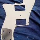 Bílá pickguard pro elektrickou kytaru s dvěma obdélníkovými výřezy. V dolní části je nalepený kovový kryt s několika otvory. Na povrchu nejsou vidite…