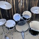 Černá bubnová sada s kovovými obručmi a stříbrnými mechanismy, doplněná o tři činely. Na činelech je vidět logo ABX Cymbals. Sada obsahuje basový bub…