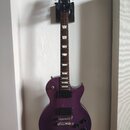 Fialová elektrická kytara s tvarem Les Paul, umístěná na nástěnním držáku. Na hlavě je viditelné zlaté logo Gibson. Kytara má dva humbuckery, čtyři o…