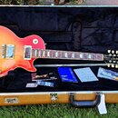 Elektrická kytara s červeným gradientním lakem (sunburst) a hnědým hmatníkem v otevřeném hnědém kufru. Na hlavě kytary je viditelné logo Gibson a náp…