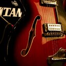 Detail polodublové elektrické kytary v červeném gradientním lakování s f-otvorem. Nástroj má dva humbuckery a kovový most. Na pozadí je vidět část če…