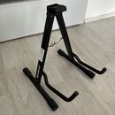 Černý kovový stojan na hudební nástroj v tvaru A. Na jedné z nohou je bílý nápis RockStand®. Povrch stojanu vykazuje mírné známky opotřebení.