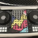 Černý DJ 컨트롤러 umístěný v přepravním kufru. Střední část přístroje je pokryta barevnou polepkou s růžovými, žlutými a modrými geometrickými tvary. V p…
