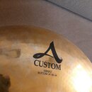 Detail zlatavého činchového činely s viditelným broušením povrchu. Na činely je černý nápis CUSTOM a nad ním stylizované logo A. Pod nápisem je uvede…
