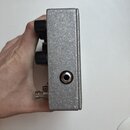 Detailní pohled na boční stranu efektového pedálu s texturovaným stříbrným povrchem. Na boku je umístěn jeden vstupní/výstupní jack konektor. V horní…
