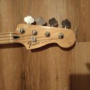 Hlava basové kytary z přírodního dřeva s четыřmi kovovými ladičkami. Na dřevě je černý nápis Fender® a PRECISION BASS®. Povrch nástroje vypadá čistě…