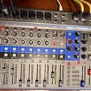Stříbrný digitální mixážní pult s nápisem L-12 LiveTrak a logem ZOOM. Zařízení má 12 vstupních kanálů s posuvníky, knoflíky pro Gain a Comp, modré tl…