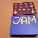 Modrá krabice s žlutým nápisem JAM a menším červeným nápisem PEDALS v pravém dolním rohu. Na krabici leží hnědý kartonový vložkový materiál s vyřezan…