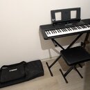 Černý digitální klavír umístěný na černém skládacím stojanu s přívodním kabelem. Na nástroji je vidět nápis YAMAHA a doprovodná hudební notová opěrka…