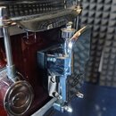 Detailní záběr na chromovaný mechanismus snare throw (přepínač čala) u červeného bubnu. Na chromovaném kovovém dílu je vyryto logo Mapex. Povrch je l…