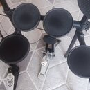 Elektronická bicí souprava s černými gumovými pady a černým rámem. Na jednom z padů je viditelné logo Roland. Součástí je kovový pedál s bílým knoflí…